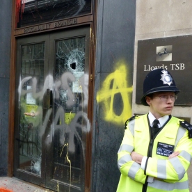 A young policeman in front of a Lloyd's of London entrance whose glass has been smashed and grafittied.