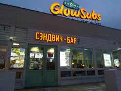 Signage in Roman and Cyrillic for Glow Subs attached to fast food restaurant with people inside