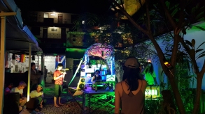 In the courtyard lit by blue and green light, band of electronic musicians and singer perform while one man films and others listen and watch
